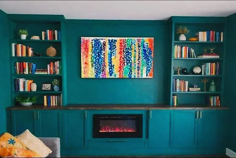 A living room with teal painted walls, two built-in bookshelves filled with books and decorative objects, a colorful abstract painting centered above a teal cabinet, and an electric fireplace below the painting.