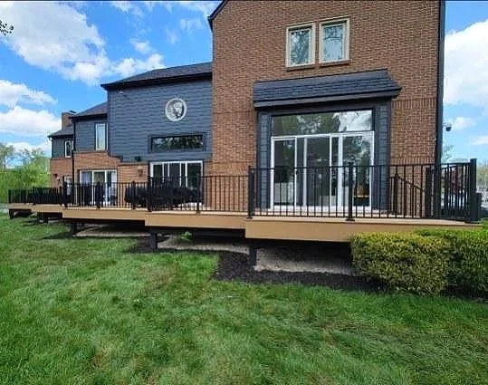 View of a backyard deck attached to a brick house with sliding glass doors, black metal railing, and neighboring houses in the background.