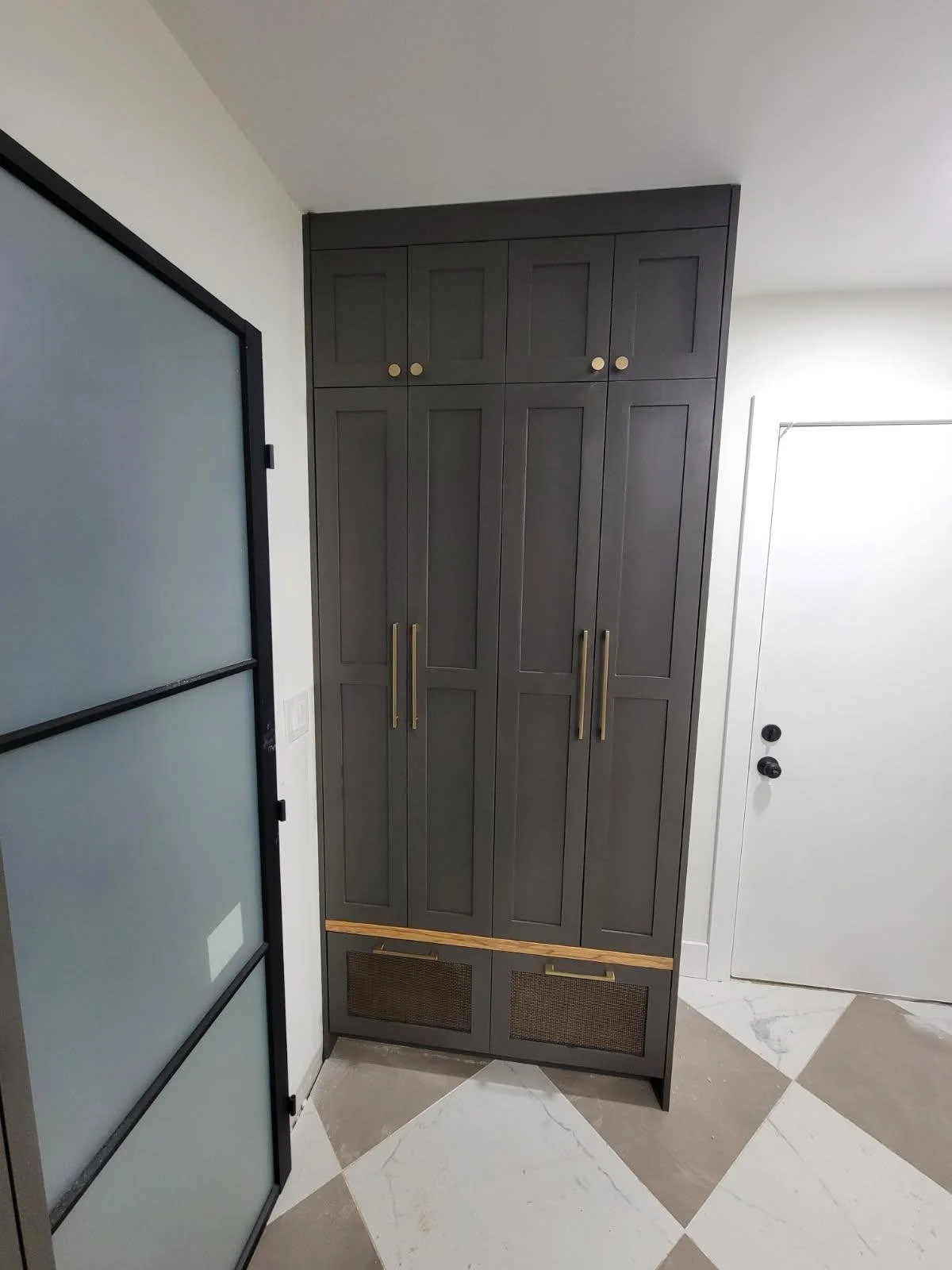 Tall dark gray cabinet with gold handles in a modern interior next to a white door and frosted glass sliding door with black frame.