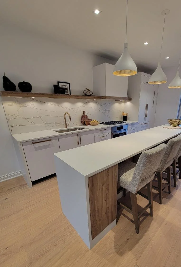 Modern kitchen with white cabinets, marble backsplash, wooden floating shelf, and a white countertop island with seating. Pendant lights hanging from the ceiling, and some decorative items on the shelf.