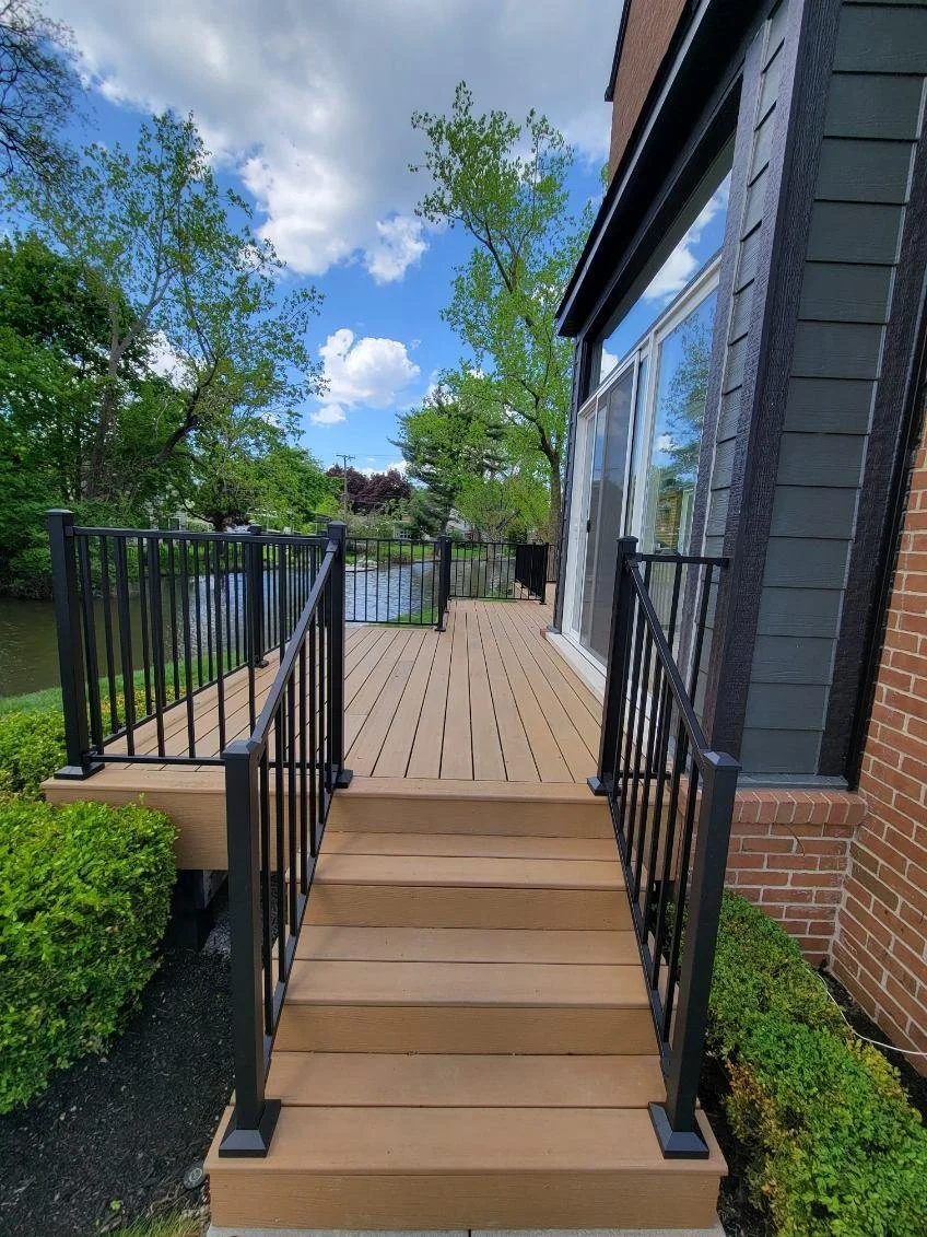 Wooden deck with black metal railings outside a house, overlooking a tree-lined pond under a partly cloudy sky.
