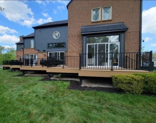 View of a modern brick townhouse with a spacious wooden deck extending from the sliding glass doors, with black railing, surrounded by green grass and bushes, under a partly cloudy sky.