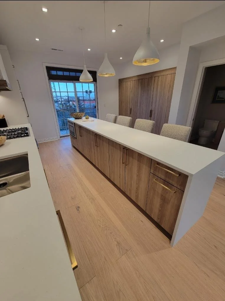 Modern kitchen with a white island, wooden cabinets, beige barstools, pendant lights, and a sliding glass door leading to a balcony with a city view.