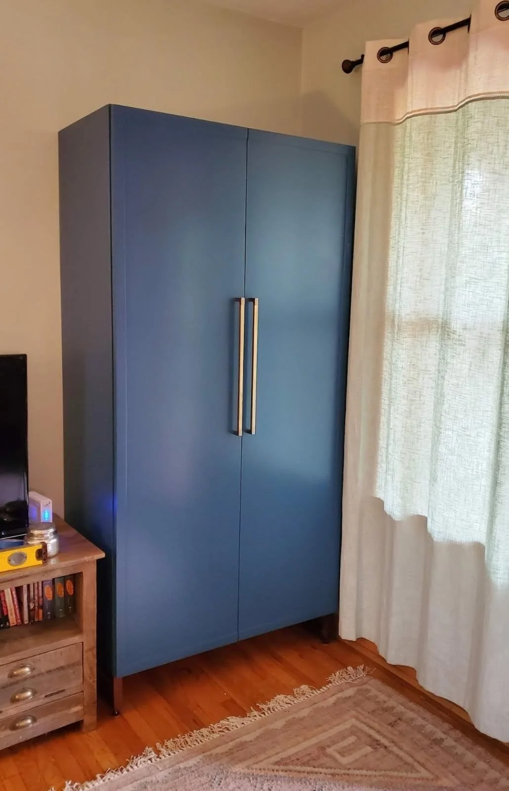 A blue wardrobe with two vertical handles next to a window with beige curtains, in a room with wooden flooring and a beige rug.