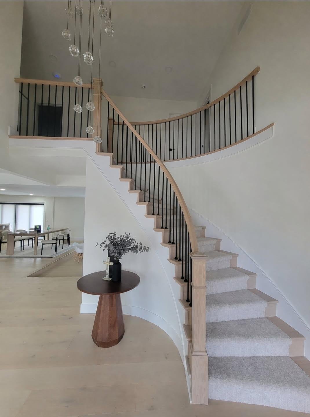 Interior view of a modern house showing a staircase with black metal balusters and wooden handrails, a hanging glass light fixture, a small round wooden table with a vase of dried flowers, and a dining area with a large wooden table and chairs in the background.