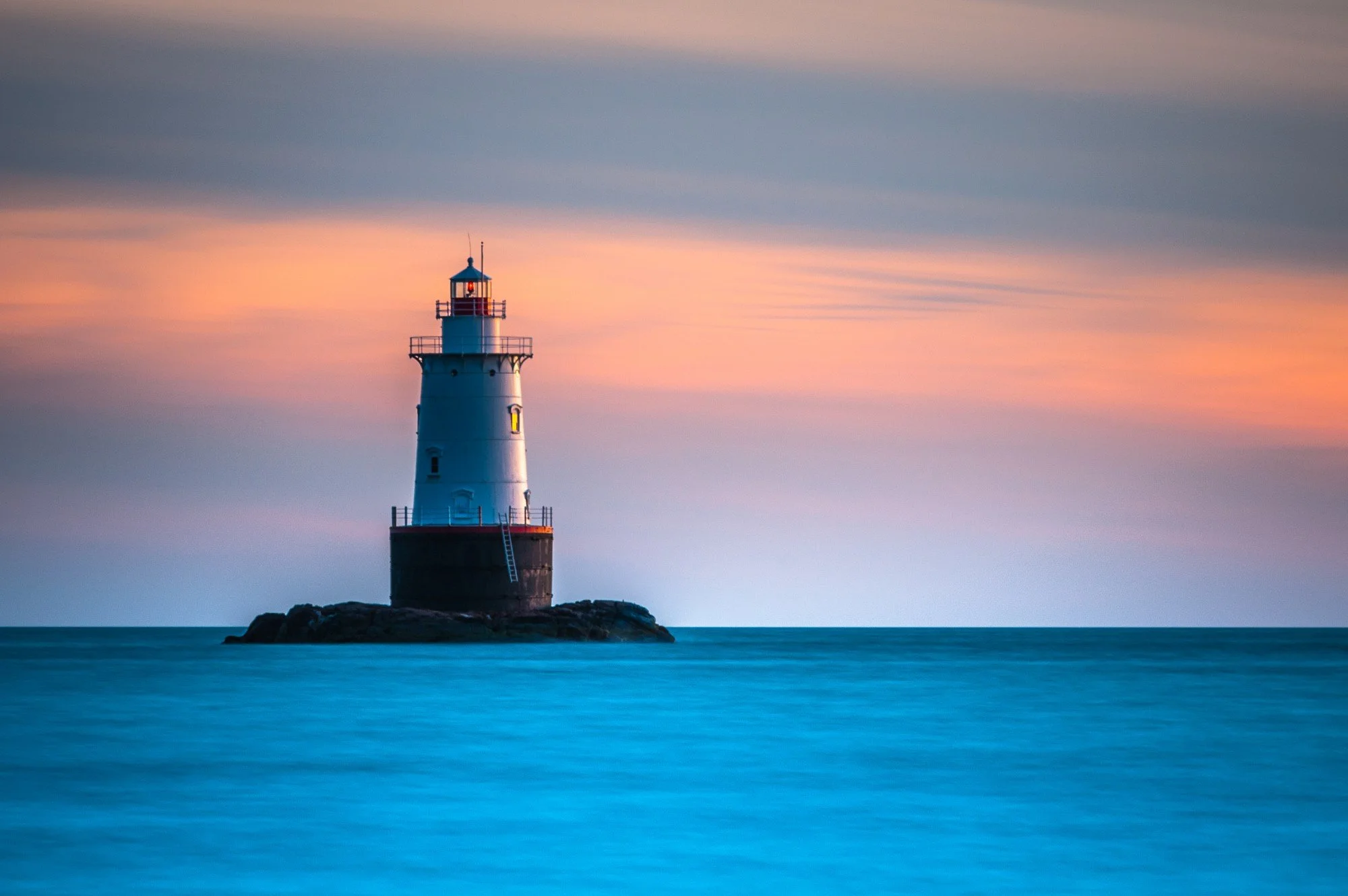Lighthouse on a small rocky island at sunset with a colorful sky and calm ocean waters in the foreground.
