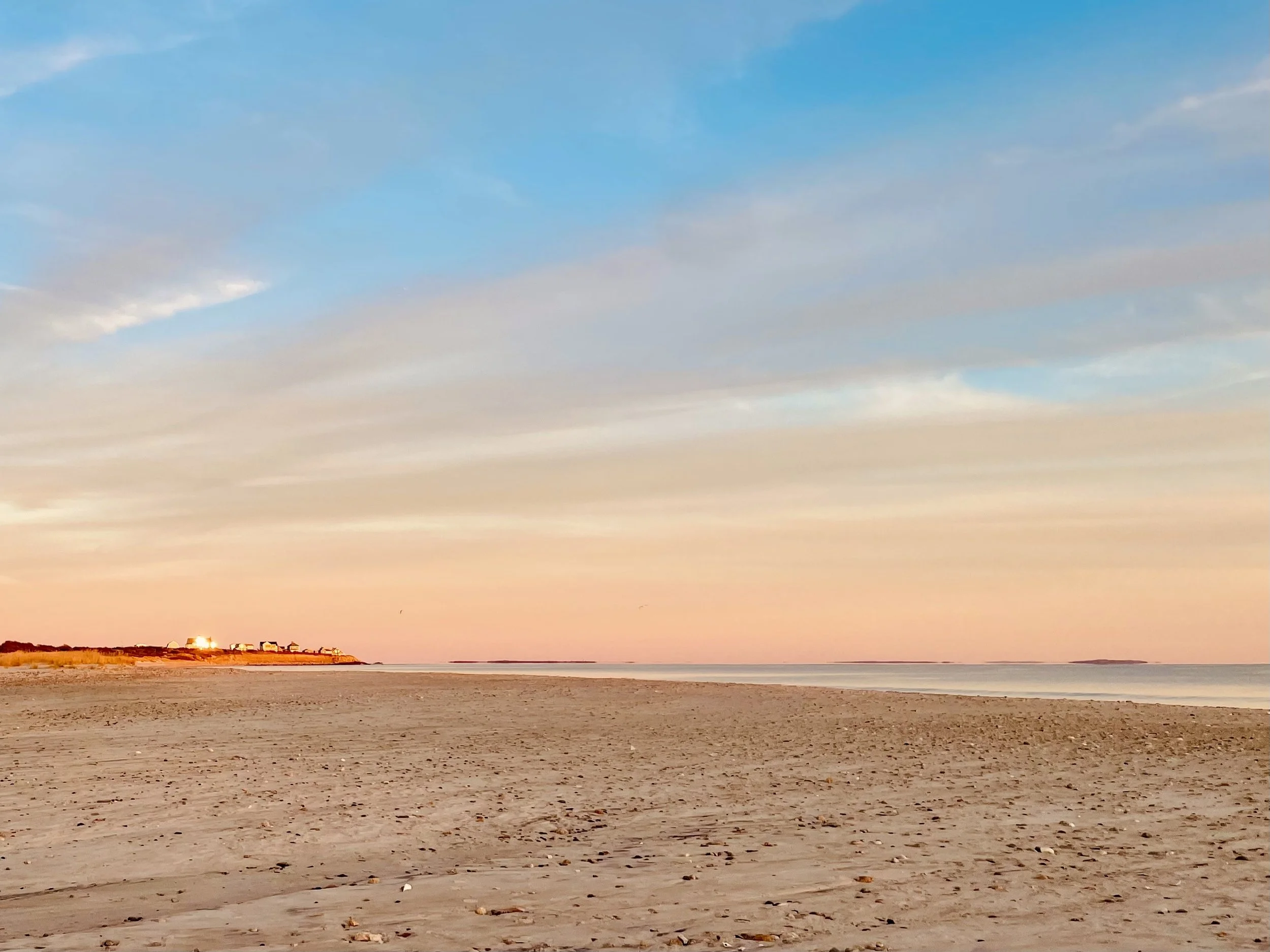 Sunset over a beach with a sandy shoreline, calm waves, and a cliff with houses in the distance, under a partly cloudy sky.