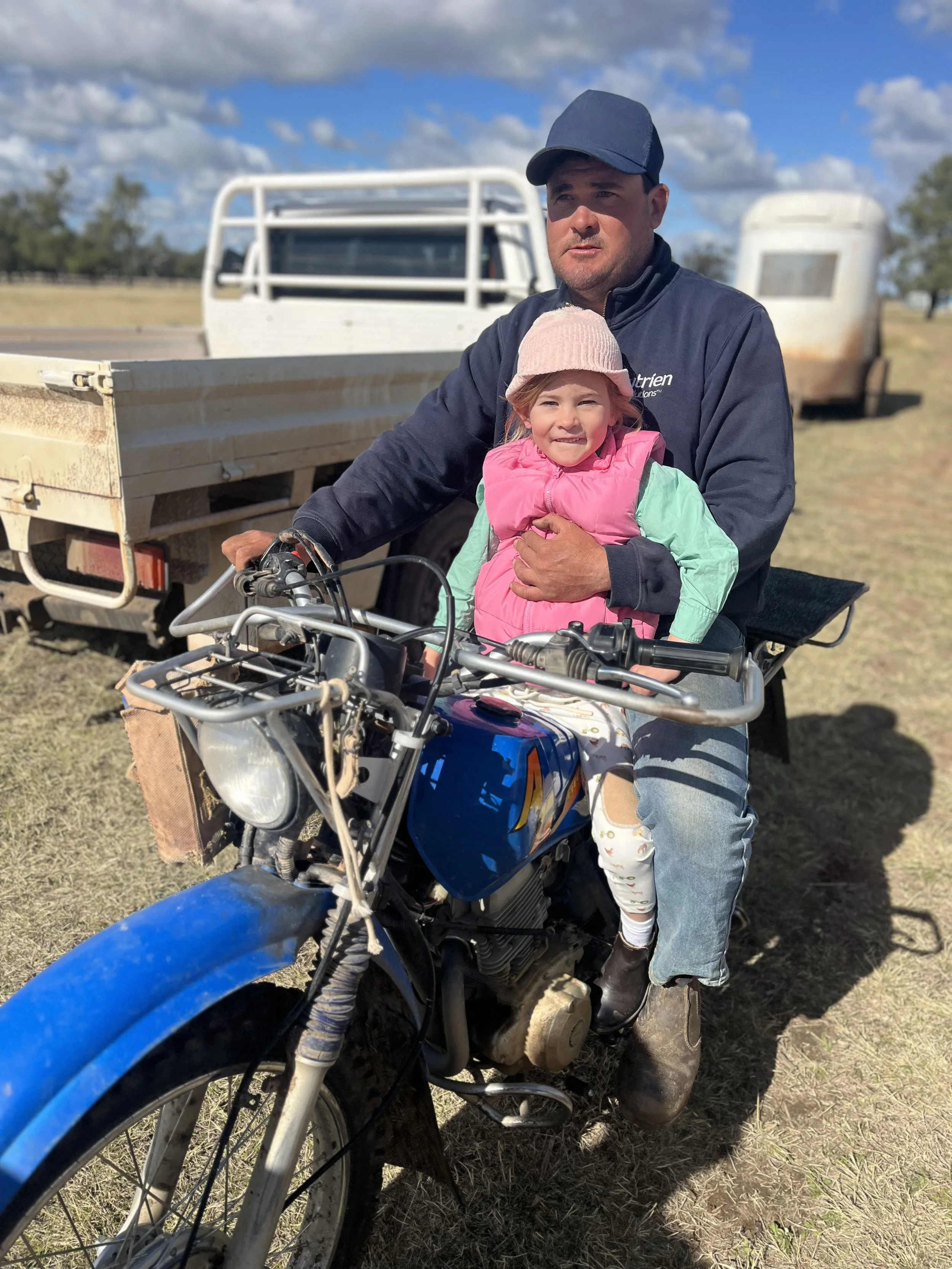 Kinsale Farms | A man and a young girl on a blue motorcycle outdoors. The man is wearing a dark jacket and cap, while the girl is dressed in a pink vest, green long-sleeve shirt, and a pink hat. 