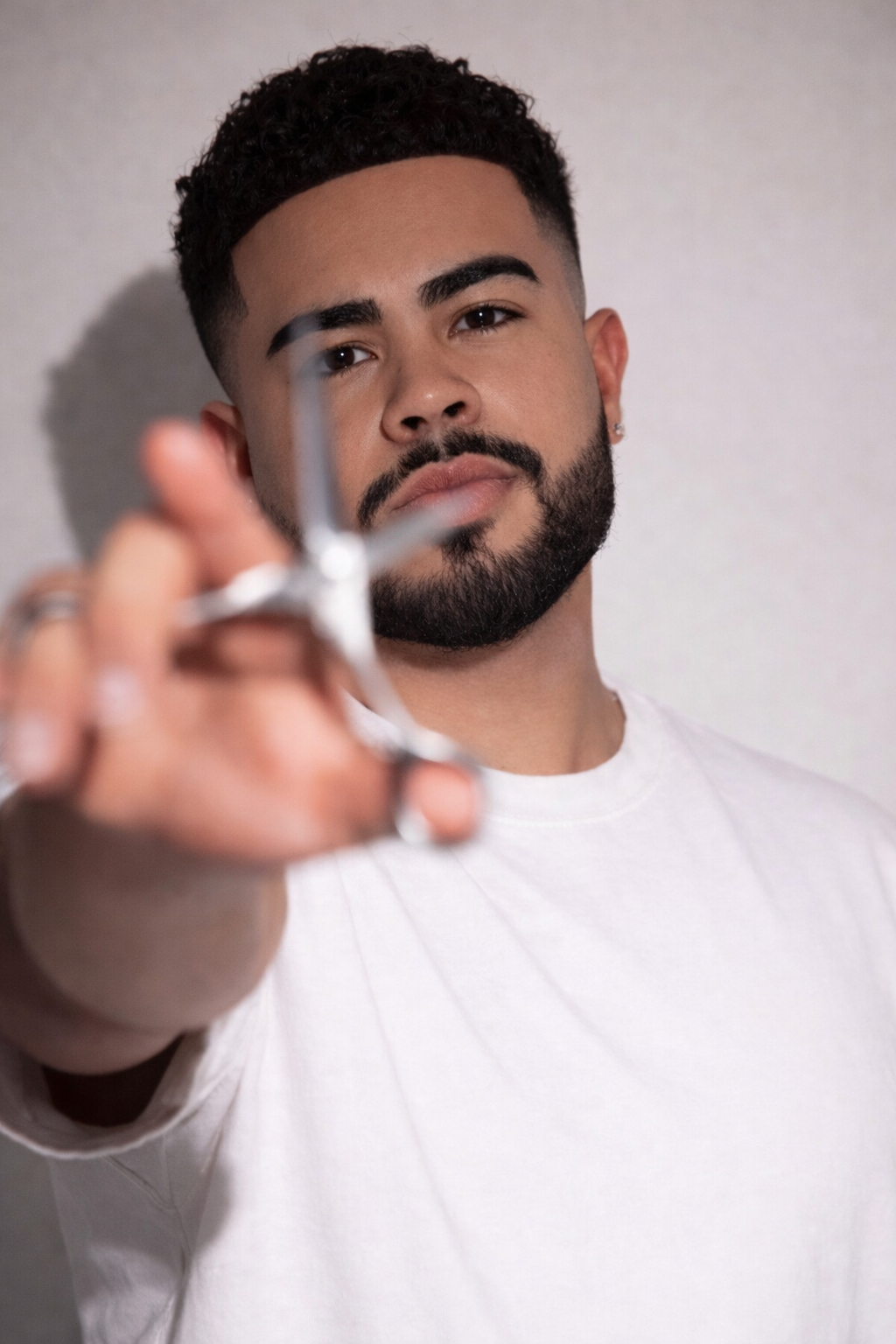A man with a beard and curly hair holding a pair of scissors toward the camera, wearing a white t-shirt, with a neutral background.