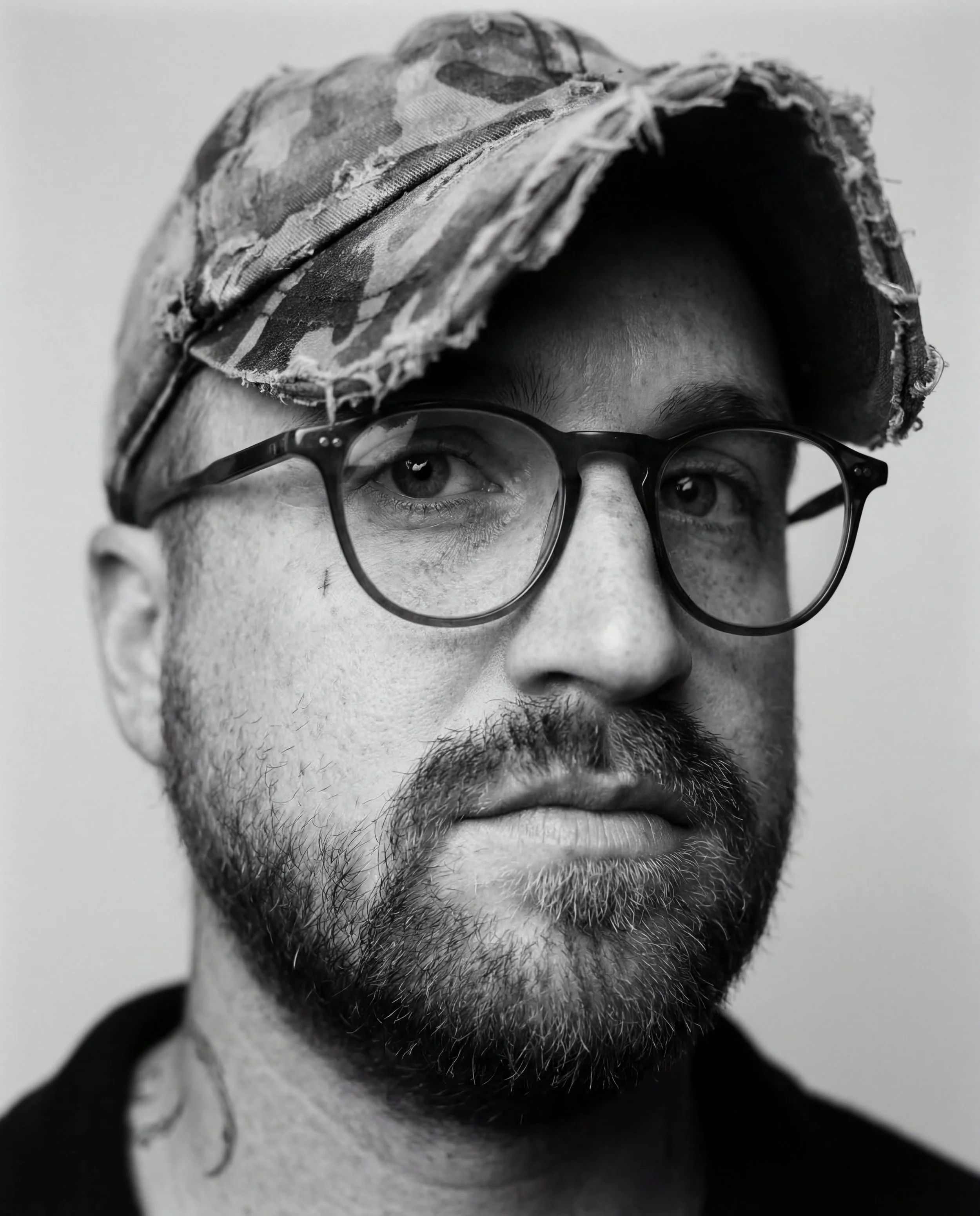 Close-up black and white portrait of a man wearing glasses and a camouflage cap, with a serious expression.