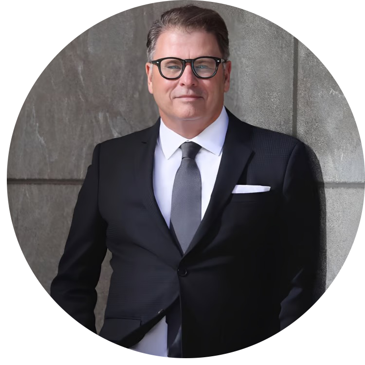 A man wearing glasses, a black suit, white shirt, and gray tie, standing against a gray stone wall, looking confidently at the camera.
