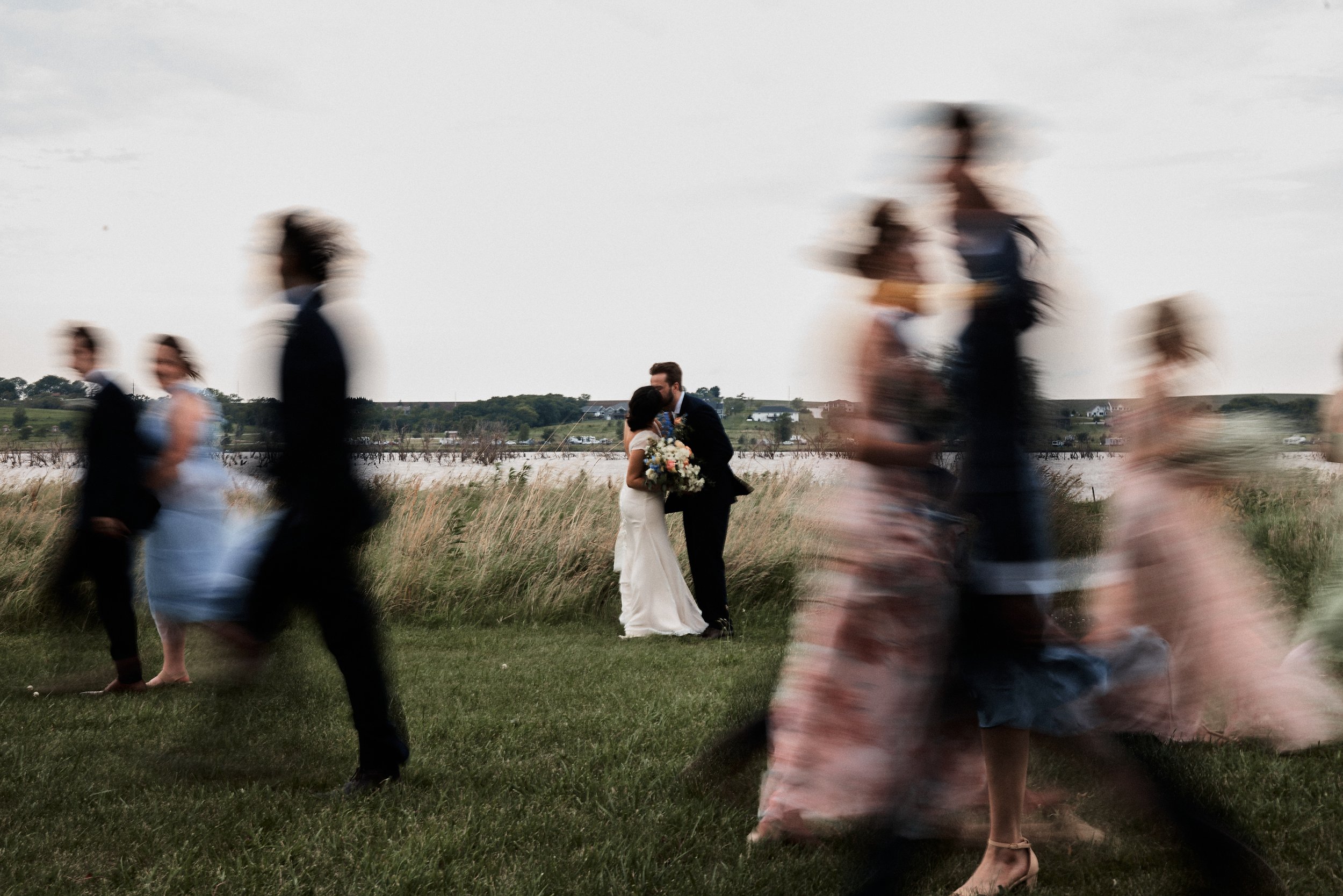 A couple, bride and groom, kissing in a field during sunset while multiple guests walk by in motion blur.