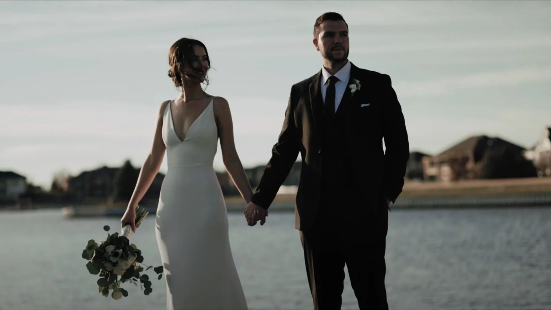 A bride and groom holding hands during their wedding by a lake at sunset, with houses in the background.