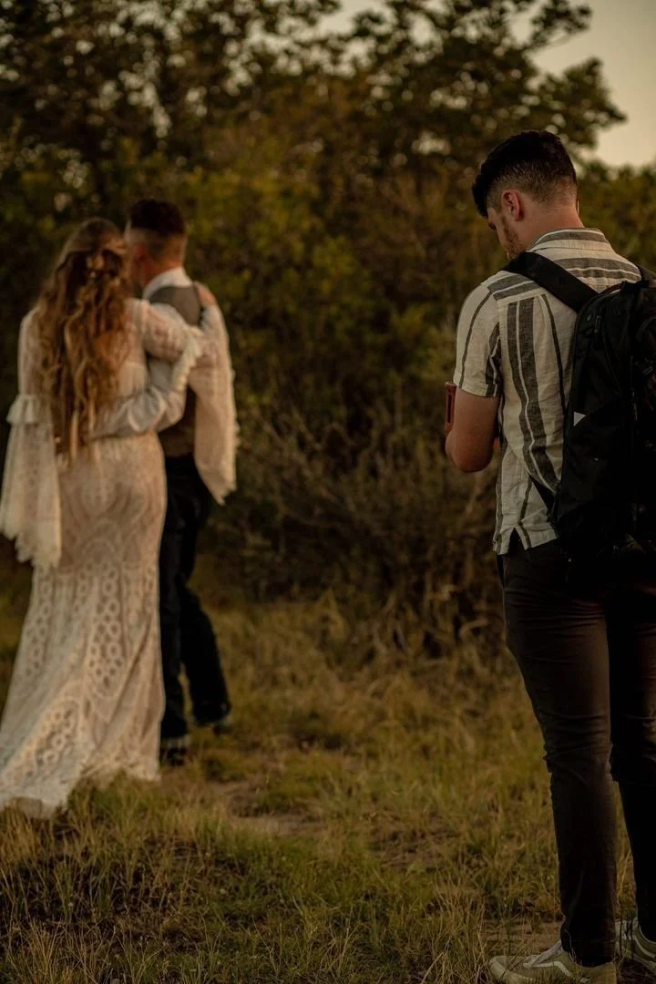 A man with a backpack photographing a couple embracing outdoors during sunset, with trees in the background.