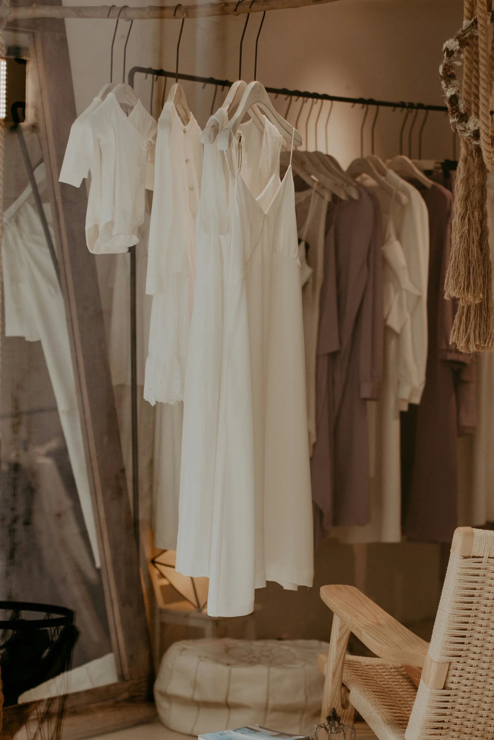 Clothes hanging on a rack with a mirror reflecting part of the room, visible chair, patterned cushion, and a magazine on a table.