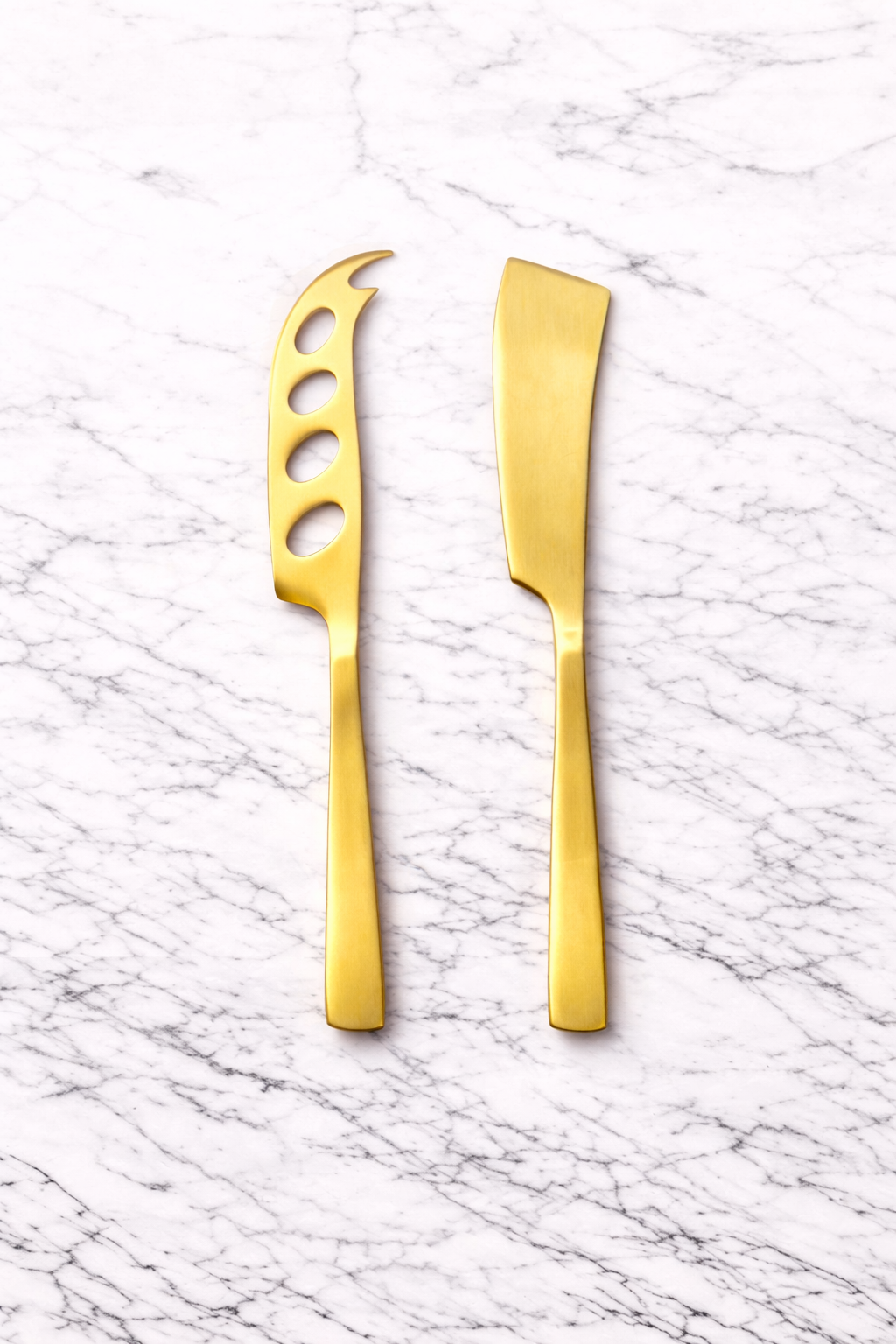 Two gold-colored cheese knives on a white marble surface. One knife has a slotted, claw-like blade, and the other has a flat, broad blade.