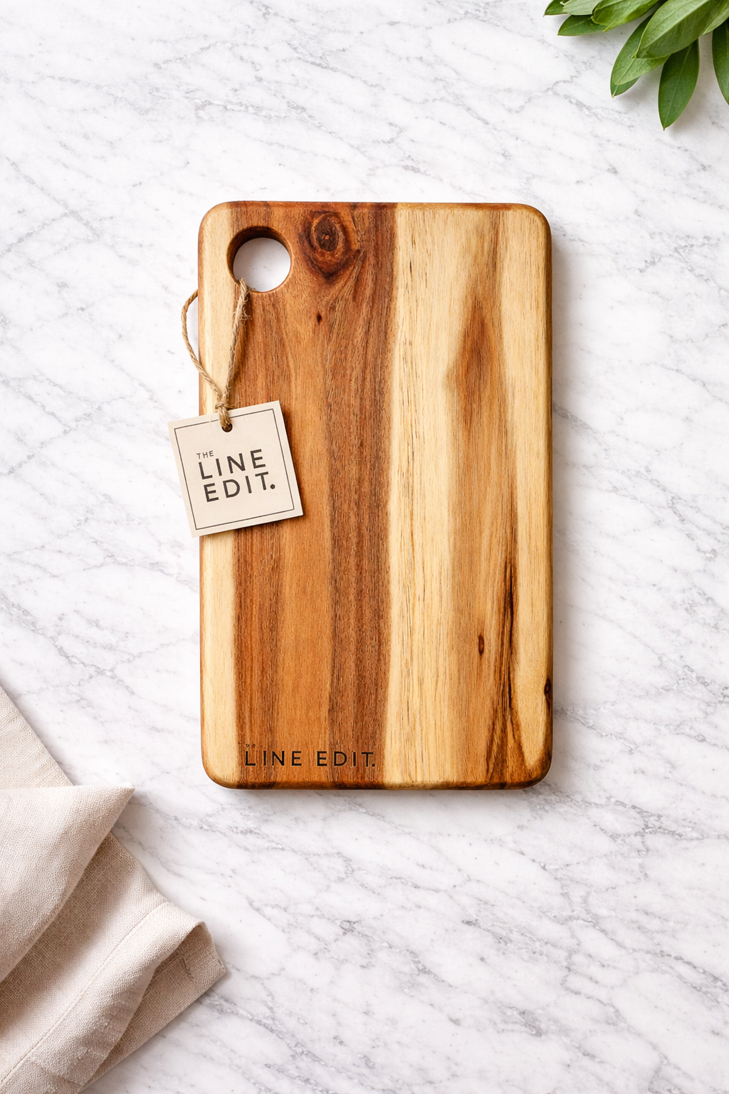 Wooden cutting board with a small tag attached reading 'The Line Edit.' on a white marble surface with a linen cloth and green leaves partially visible.