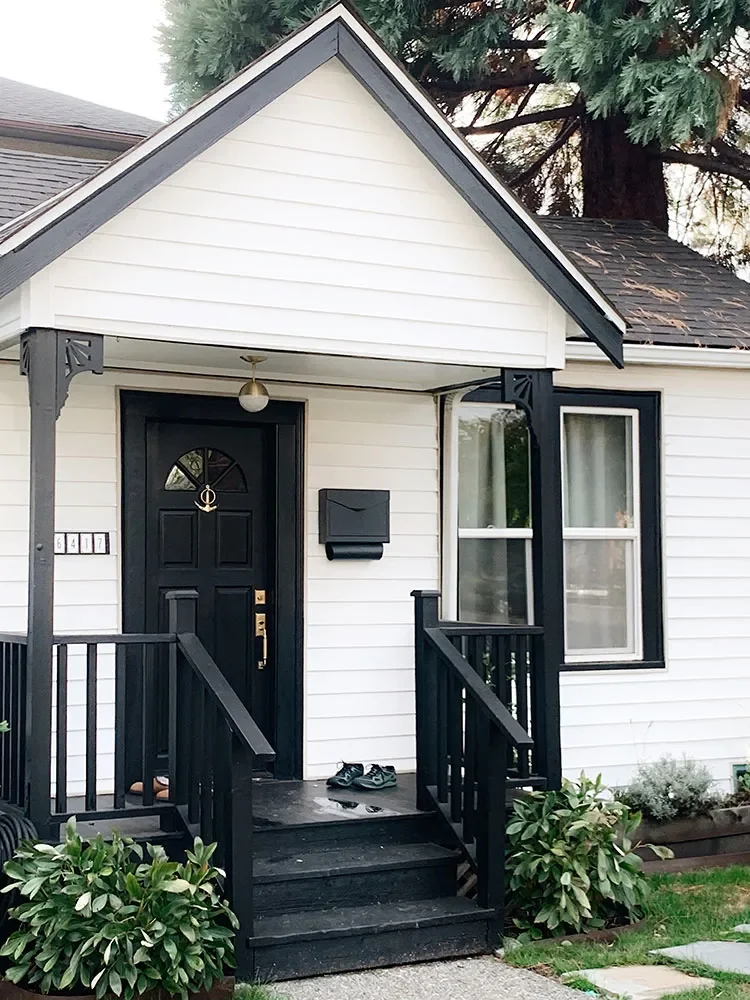 Front porch of a white house with black trim, black front door, and a mailbox. There are potted plants and shoes on the steps.