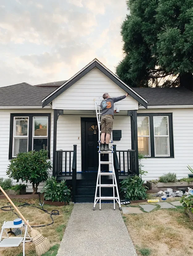 A man painting the upper part of a house’s exterior wall using a ladder, standing on the ladder, with a bucket of paint in his hand.