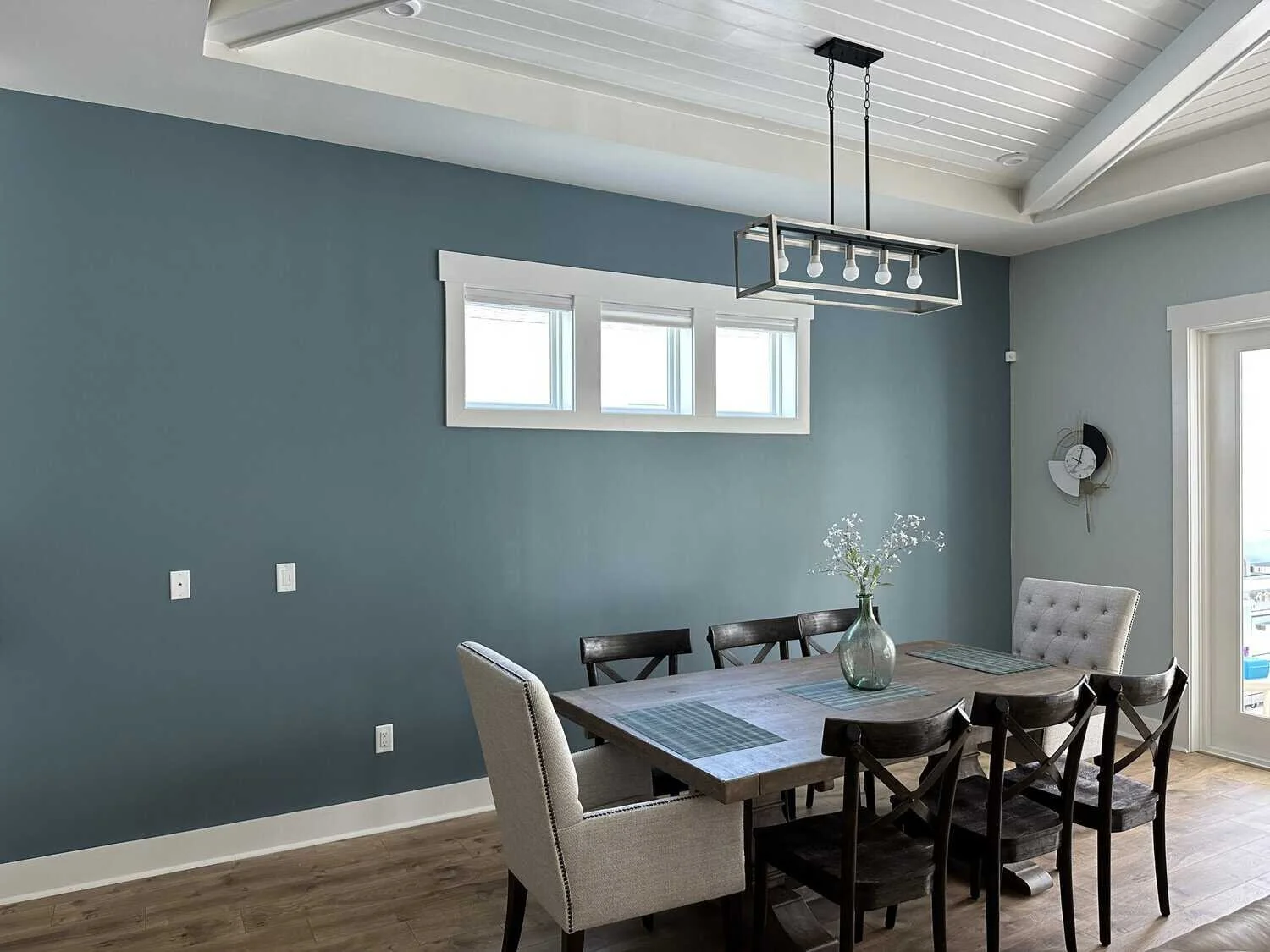 A dining room with a wooden table, six chairs, a vase with flowers, a modern pendant light, small windows, and a sliding glass door leading outside.