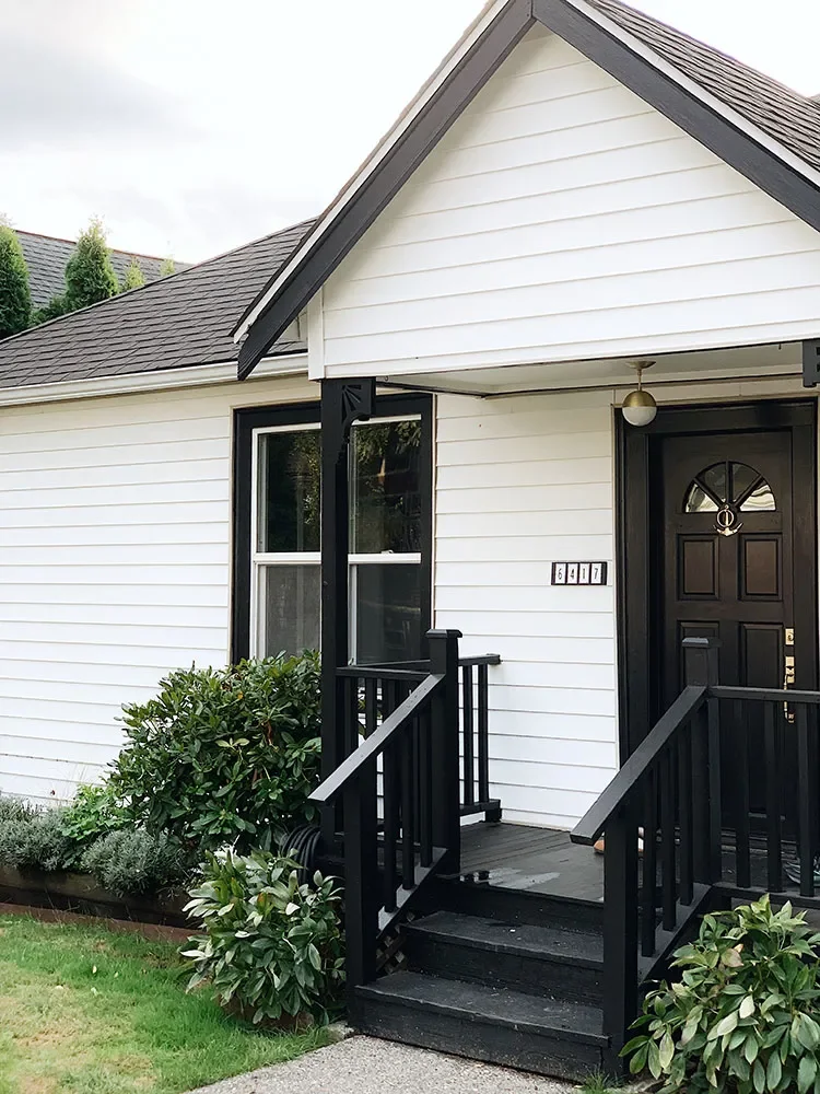 Front porch of a white house with black trim, a black door, and steps leading up to the entrance, surrounded by green bushes.
