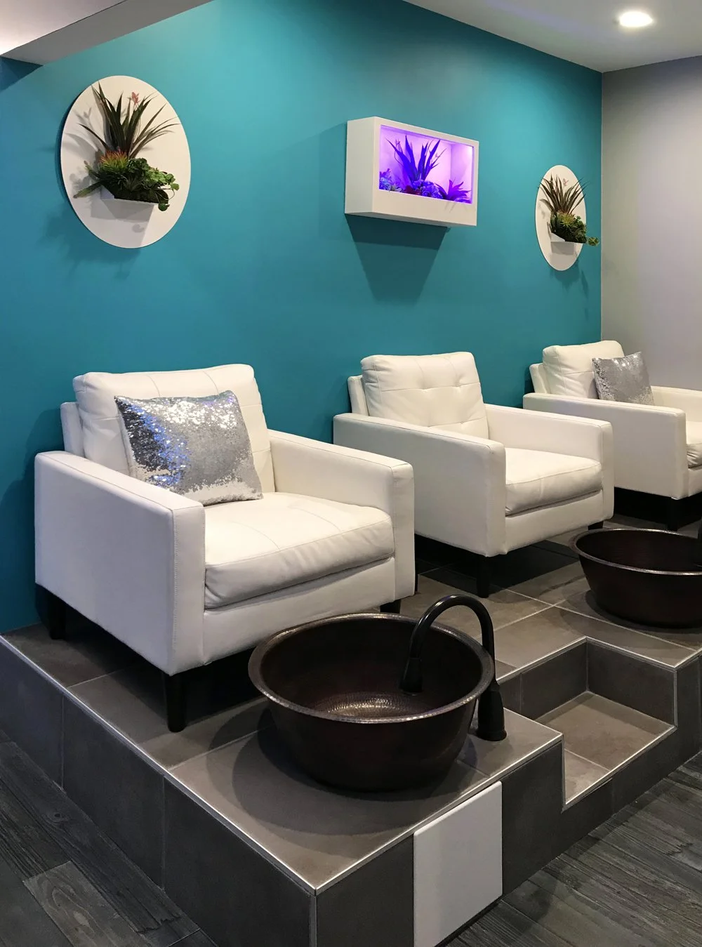 Three white salon chairs with sequin pillows in front of a teal wall with two circular wall-mounted planters and a rectangular tank with purple lighting. Two black foot baths are positioned in front of each chair.