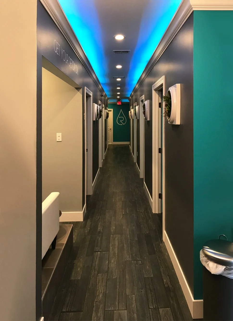 A modern, well-lit hallway in a building with dark hardwood floors, wall-mounted planters, and teal-colored walls at the end with a logo. Recessed ceiling lights and decorative crown molding are visible.