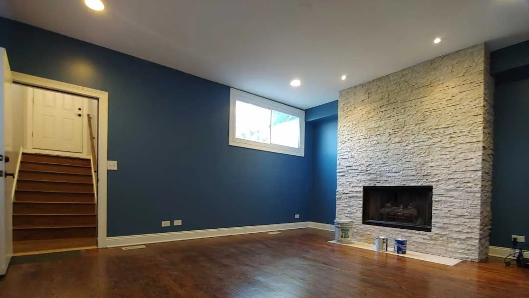 Empty living room with blue walls, hardwood floors, a white stone fireplace, a window, and stairs leading up.