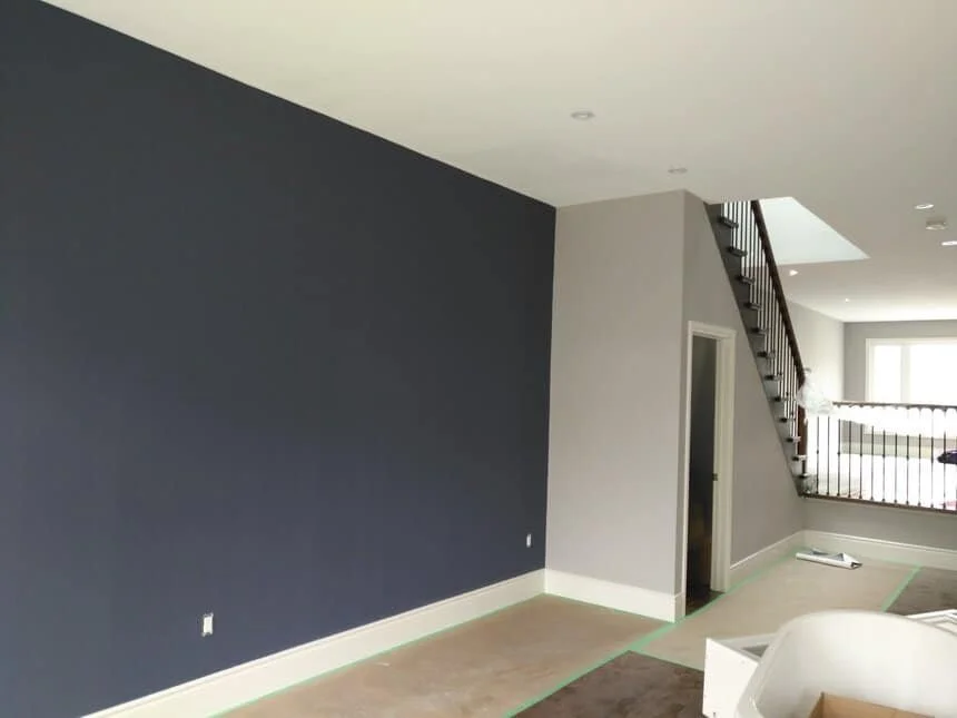 Interior room undergoing renovation with one dark blue accent wall, light grey walls, and a staircase with black railings leading to an upper floor.