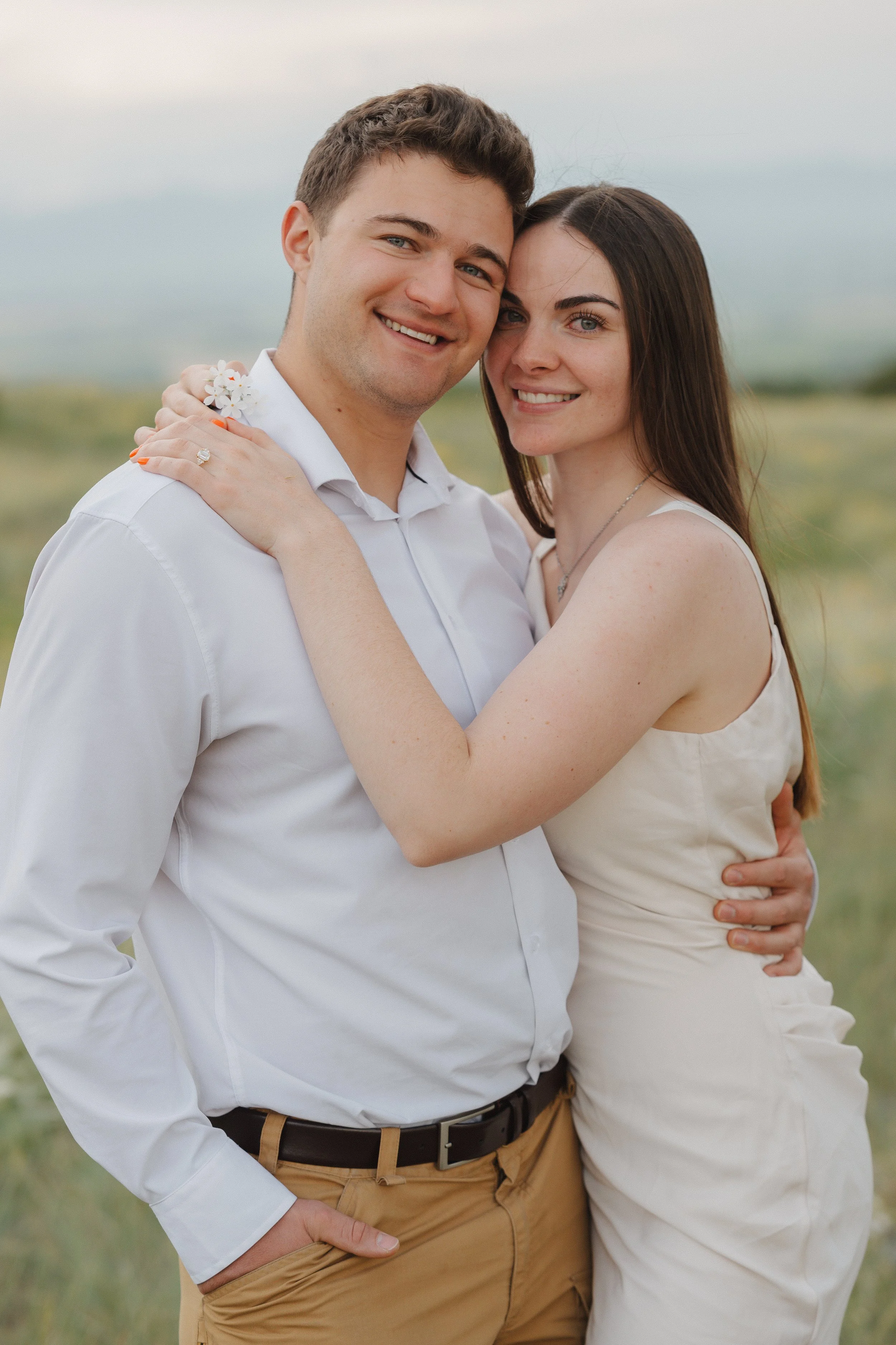 A smiling young couple embracing outdoors in a field with a soft-focus background, wearing light-colored clothing.
