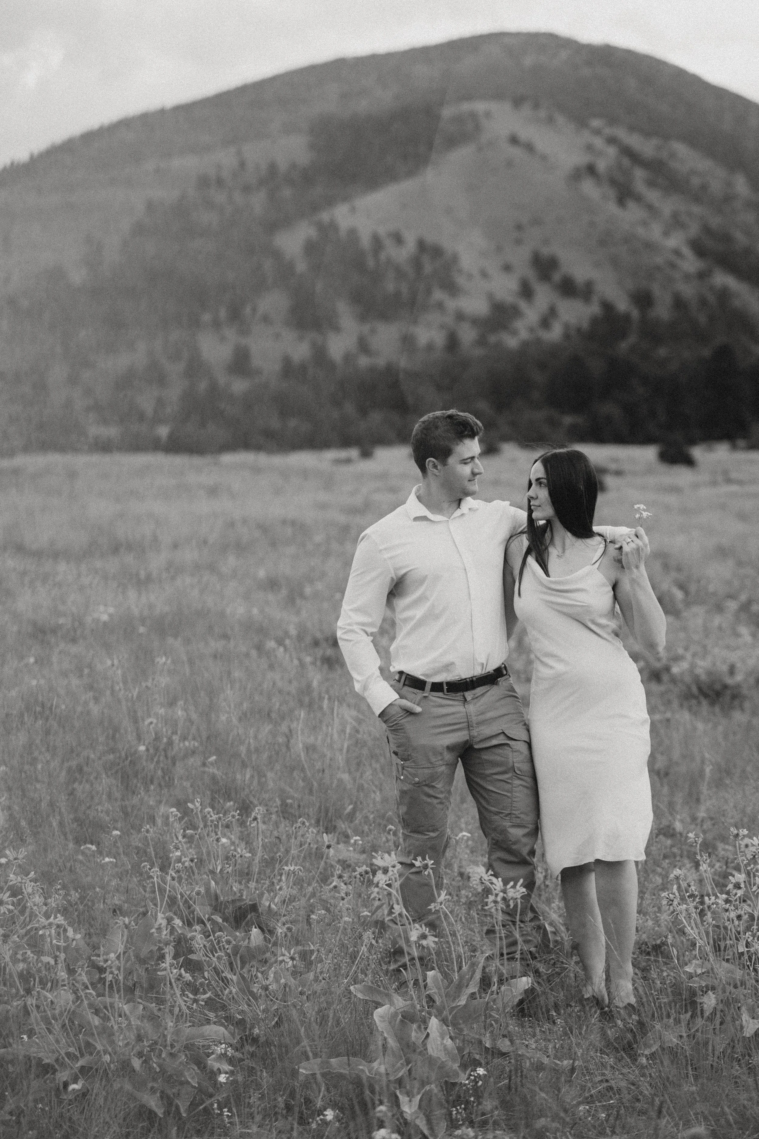 A black-and-white photo of a young man and woman walking in a field with mountains in the background. The man has his arm around the woman, who is holding a small flower.