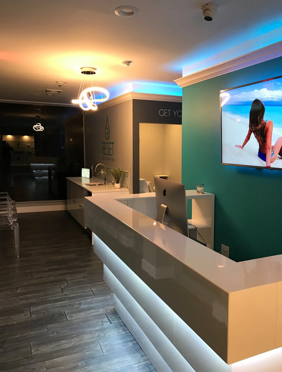 Modern reception area with sleek white counter, computer, and wall-mounted TV displaying a woman on sandy beach, teal wall, and ambient lighting.