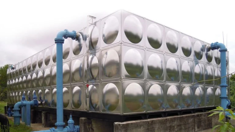 Large industrial water storage tank with a reflective metal surface and blue pipes on a concrete platform.