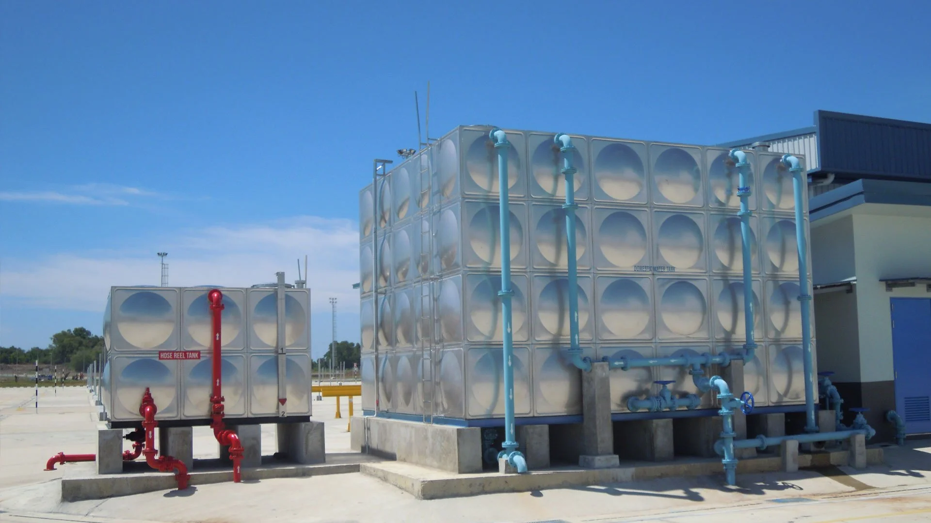 Industrial water tanks with red and blue pipes outdoors on a concrete platform against a blue sky.