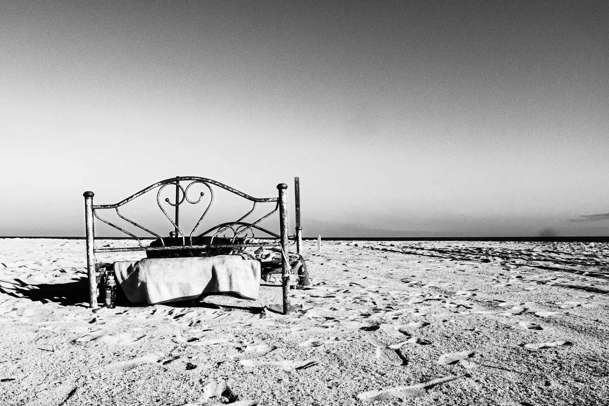Una cama aparece en medio de la playa, fuera de lugar, casi absurda a primera vista. En blanco y negro, la escena pierde lo anecdótico y gana significado: líneas simples, sombras duras y un horizonte limpio que refuerza el contraste entre objeto y en