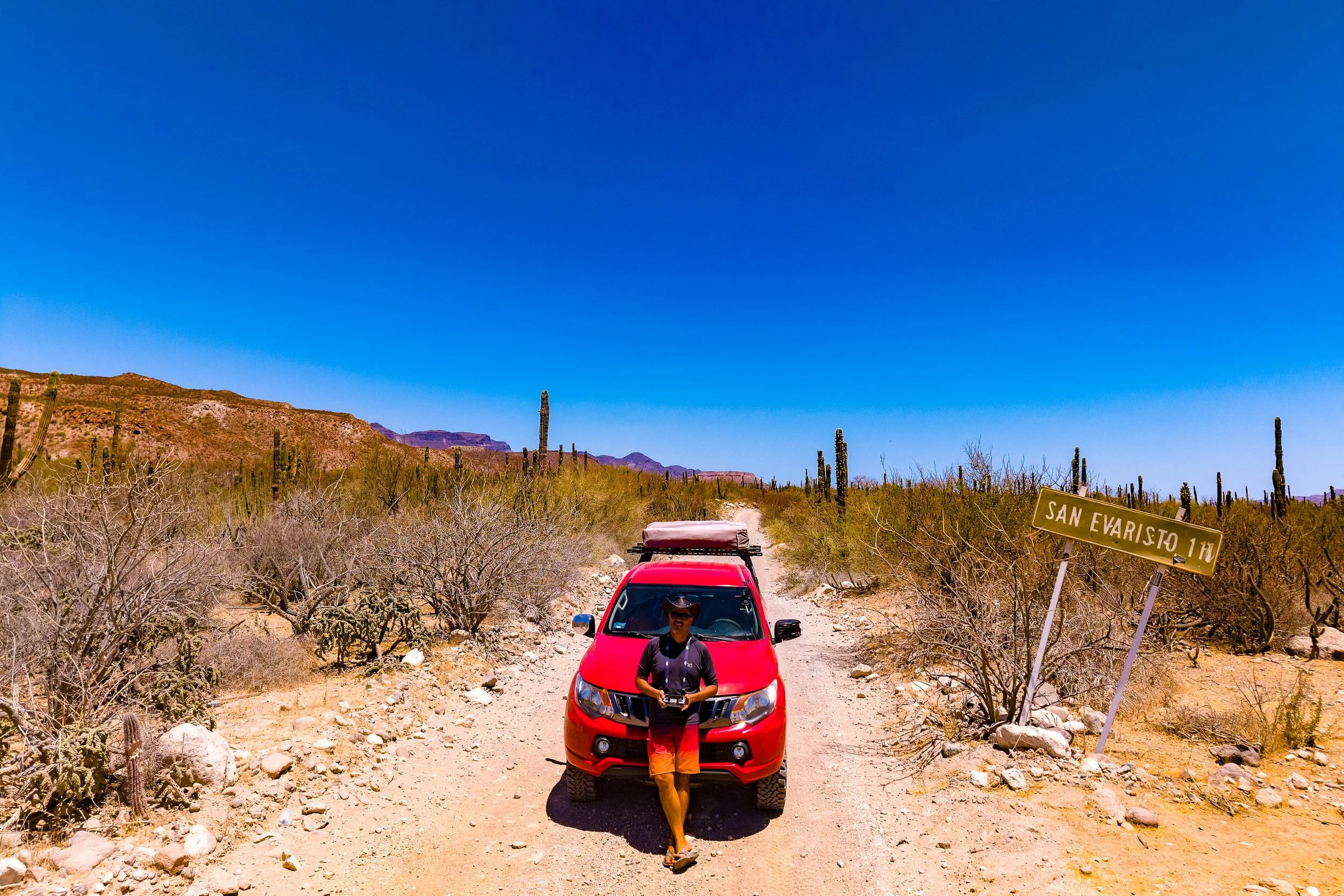 Persona con sombrero y gafas de sol de pie frente a un coche rojo en un camino en un desierto con cactus y montañas, señal de tránsito que dice 'San Evaristo 1h'.