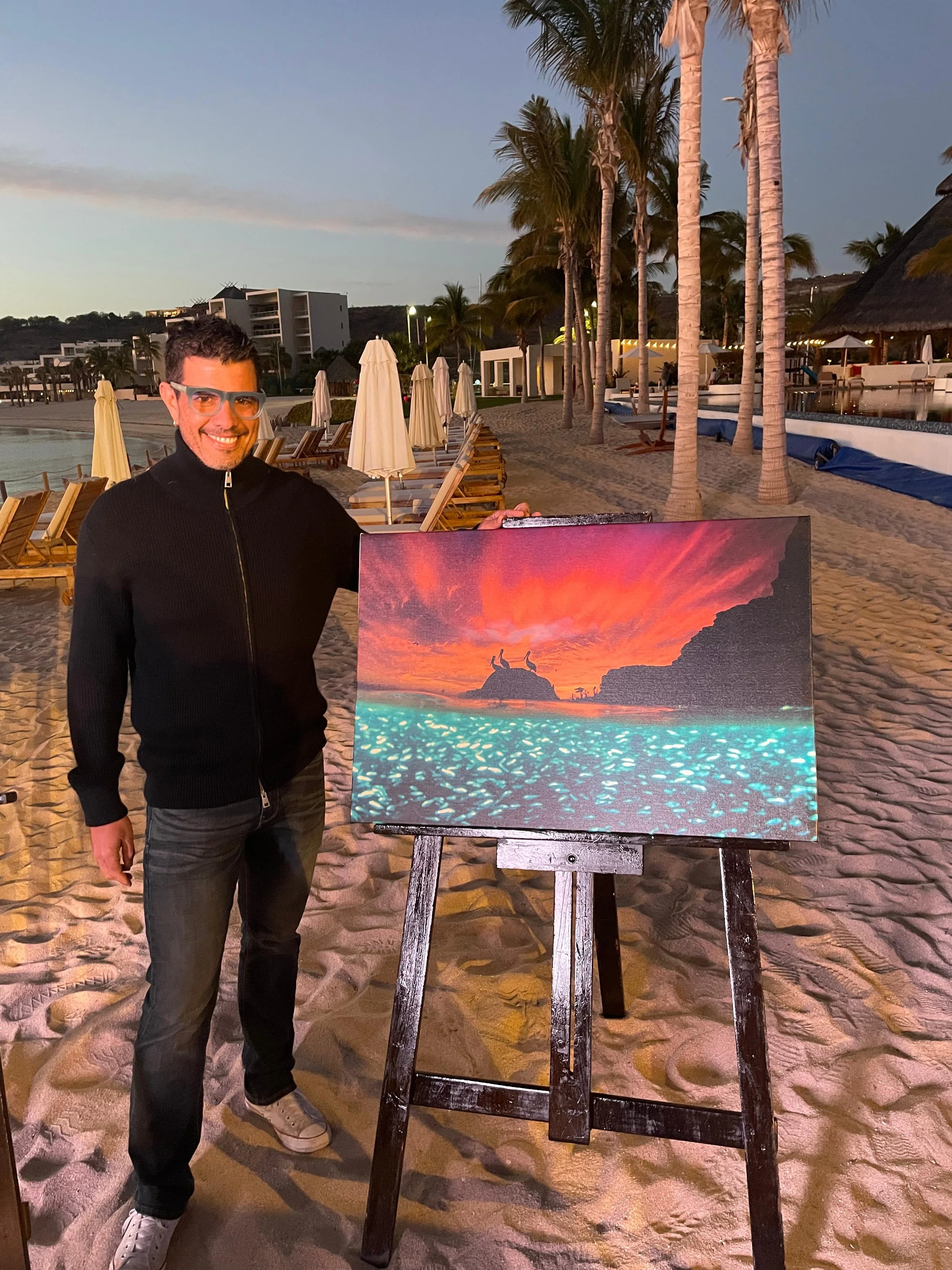 Hombre en la playa frente a un cuadro que representa una puesta de sol con el mar iluminado y formaciones rocosas.