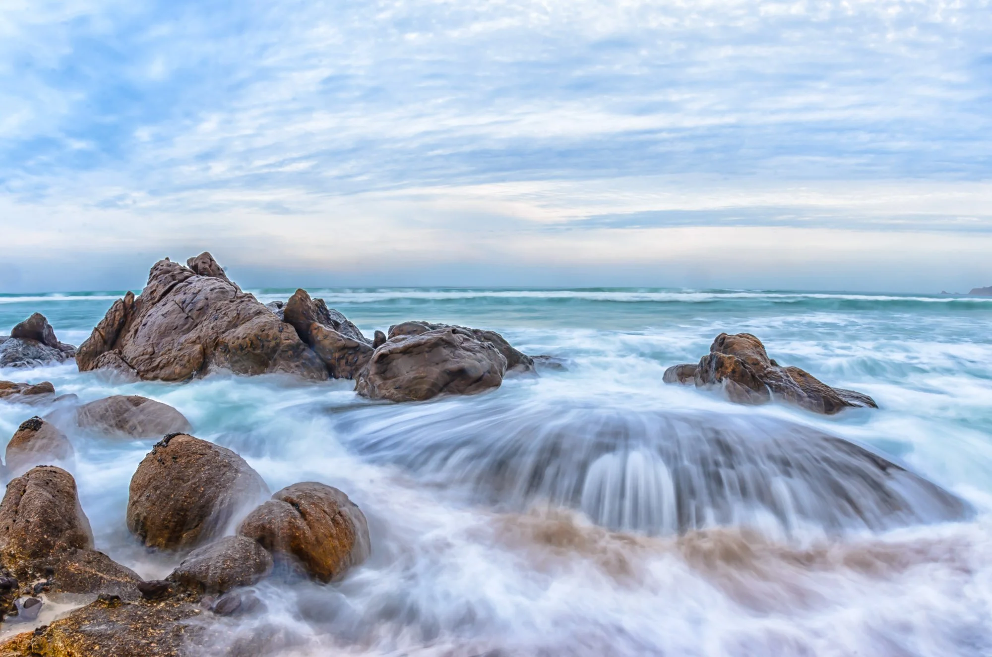 El mar golpea con fuerza una costa rocosa, pero en larga exposición esa violencia se transforma en algo casi hipnótico. El agua deja de ser caos y se vuelve flujo: líneas suaves que envuelven las rocas, espuma convertida en niebla blanca que parece d