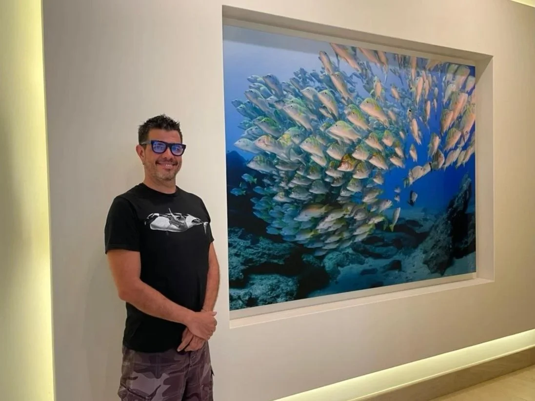 Hombre sonriendo con gafas oscuras de pie junto a una pantalla que muestra una imagen de un banco de peces en el océano.