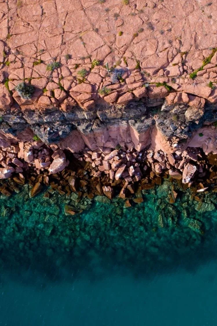 Vista desde arriba de una costa rocosa con agua cristalina en tonalidades verdes y un acantilado de rocas y tierra en su topografía.