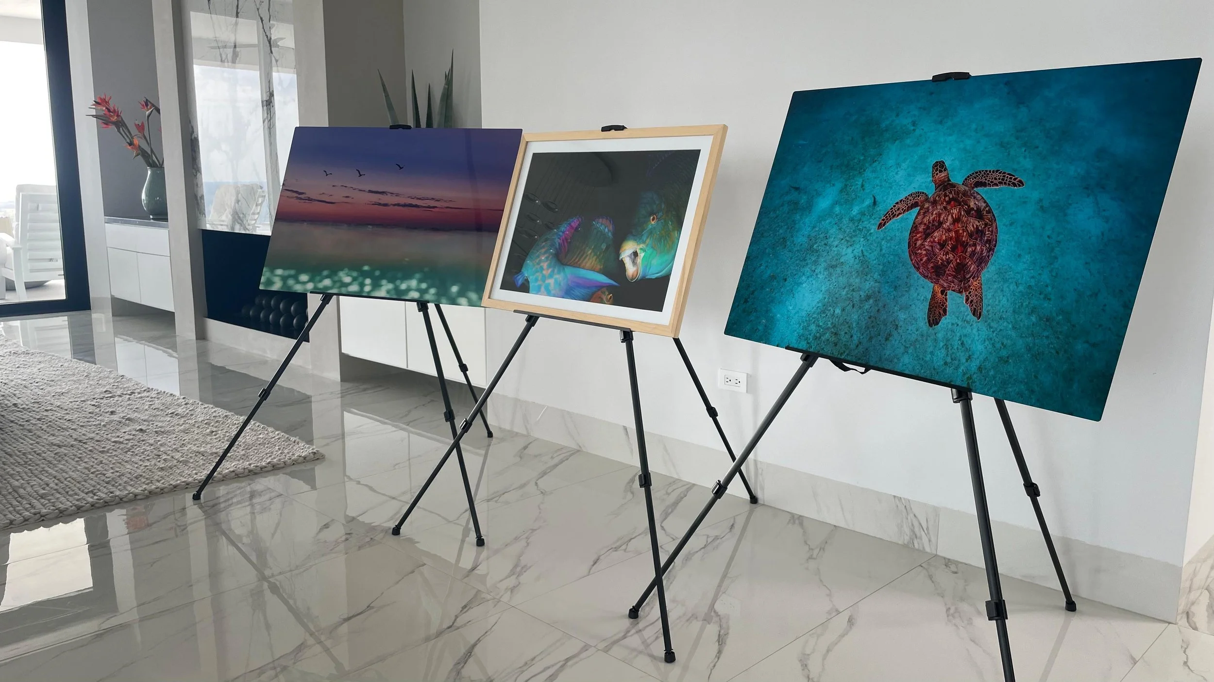 Cuatro cuadros de arte en caballetes exhibidos en una sala moderna con suelo de mármol y ventanas grandes con vista al exterior.