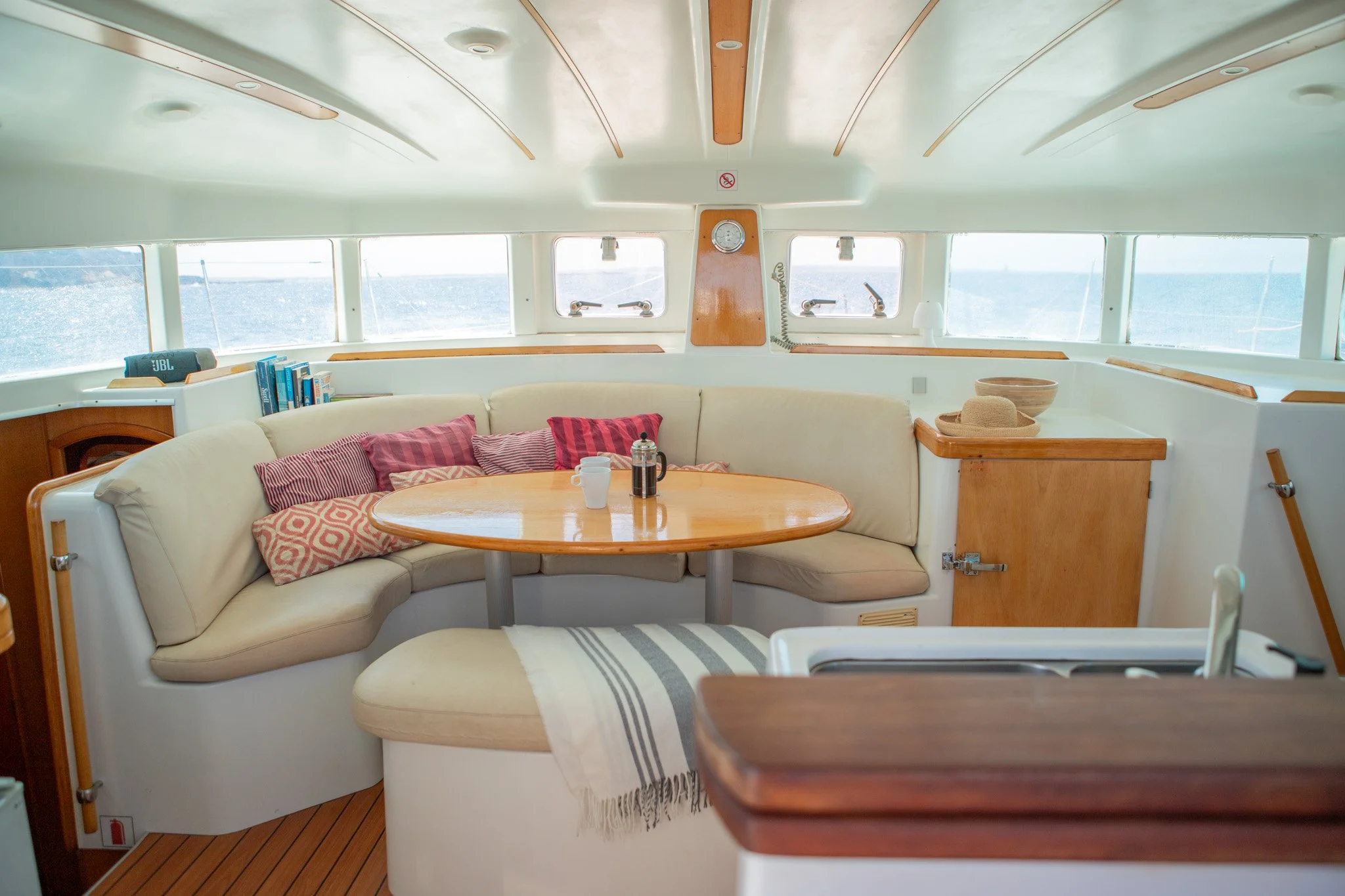 Interiores de una cabina de barco con una sala en forma de L con cojines de colores y una mesa de madera redonda. Ventanas que muestran el mar y el cielo. Decoración en madera y accesorios para beber.