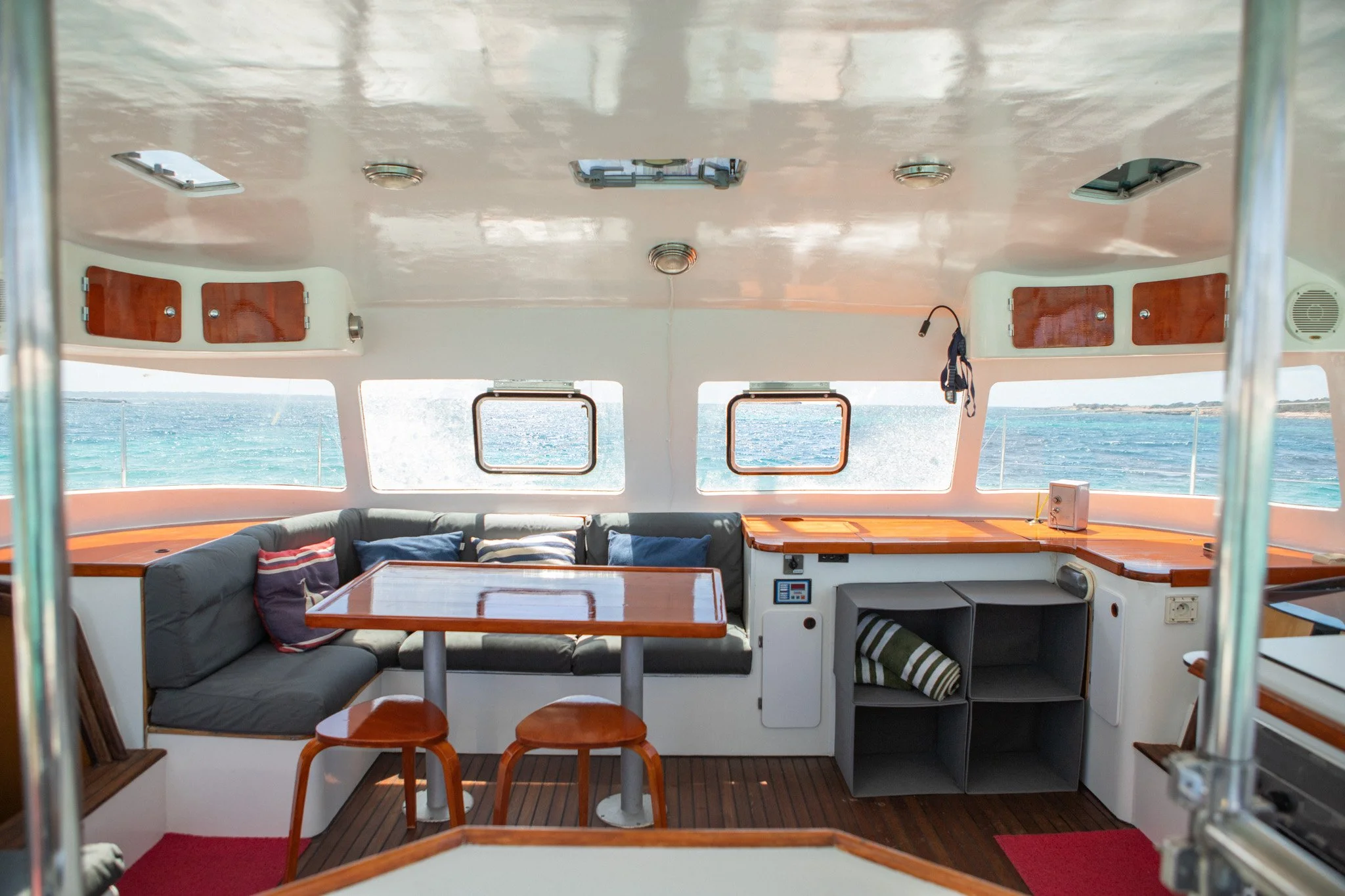 Interior de una cabina de barco con ventanas que muestran el mar y el horizonte, muebles de madera, un sofá con cojines, una mesa, y equipo eléctrico en la pared.