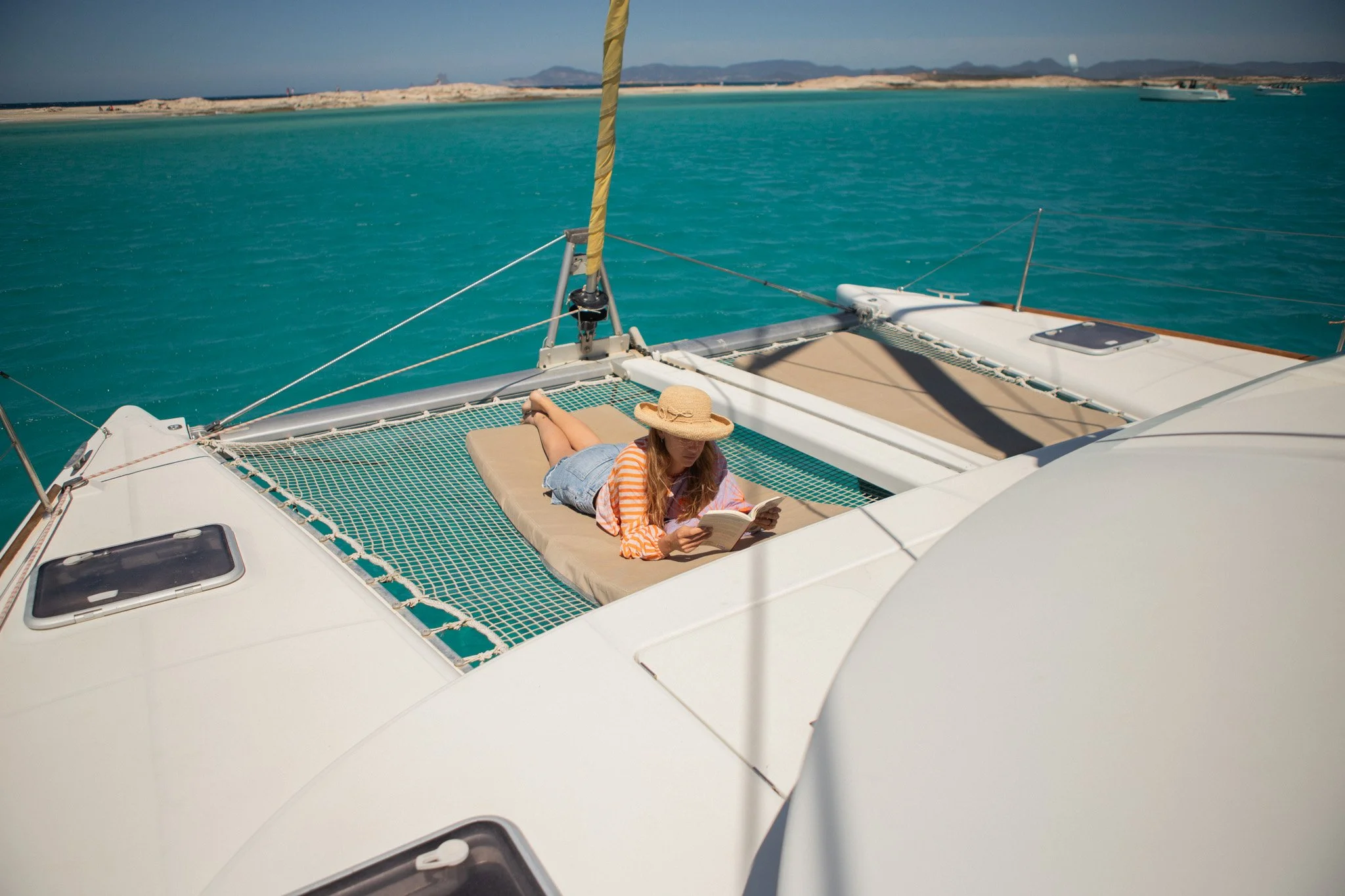 Mujer acostada en una cama en la cubierta de un barco, leyendo un libro con sombrero de paja, rodeada de agua turquesa y un cielo despejado.