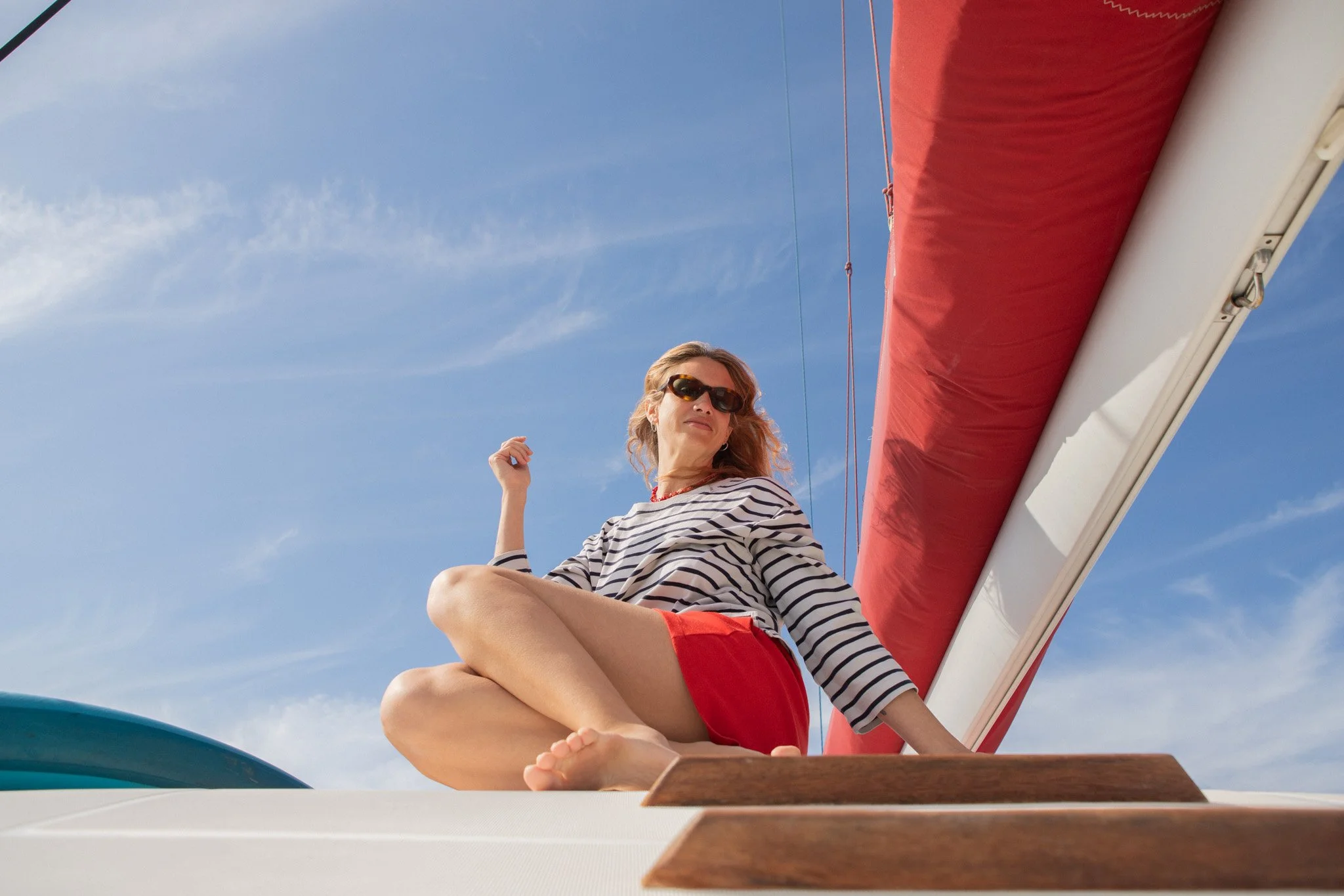 Mujer con gafas de sol sentada en un barco, con la pierna cruzada, bajo un cielo azul con nubes finas.