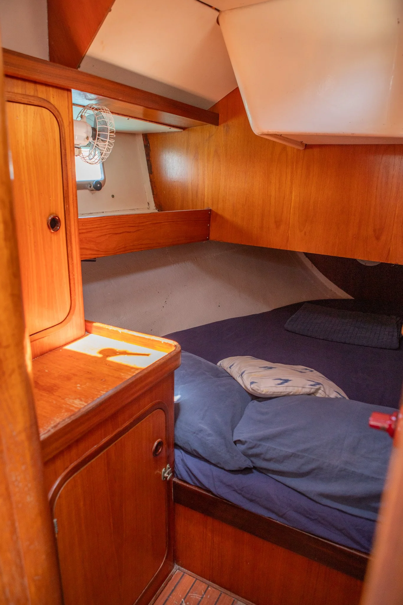 Cama con colcha azul y almohada en una habitación con paredes y mobiliario de madera, ventilador pequeño en la pared y ventana.