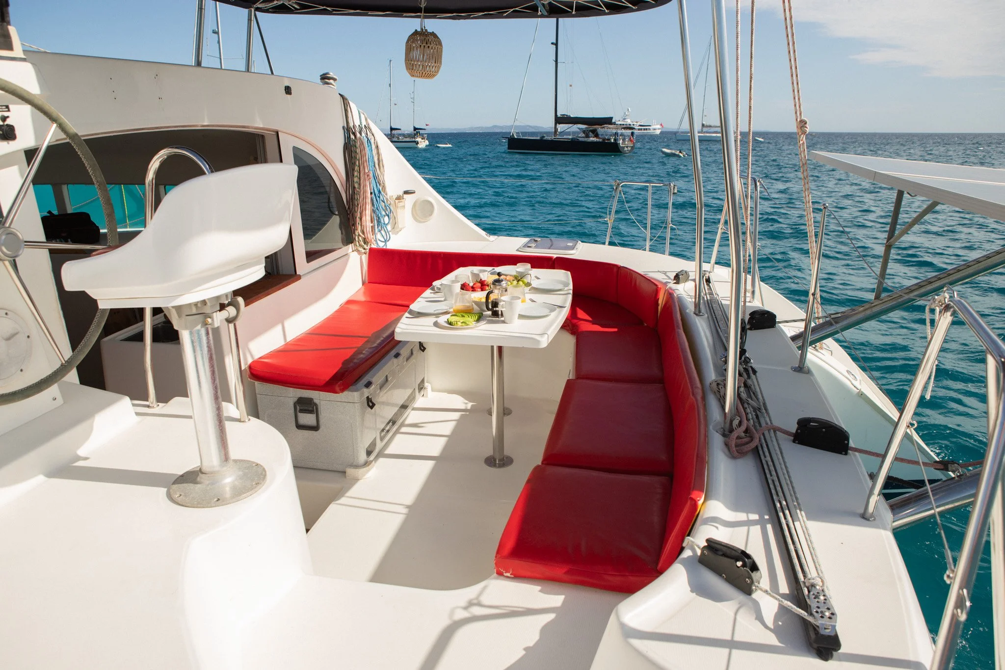 Vista del interior de un yate con mesa puesta con comida y bebidas, asientos con cojines rojos y vista del mar con otros barcos a lo lejos.