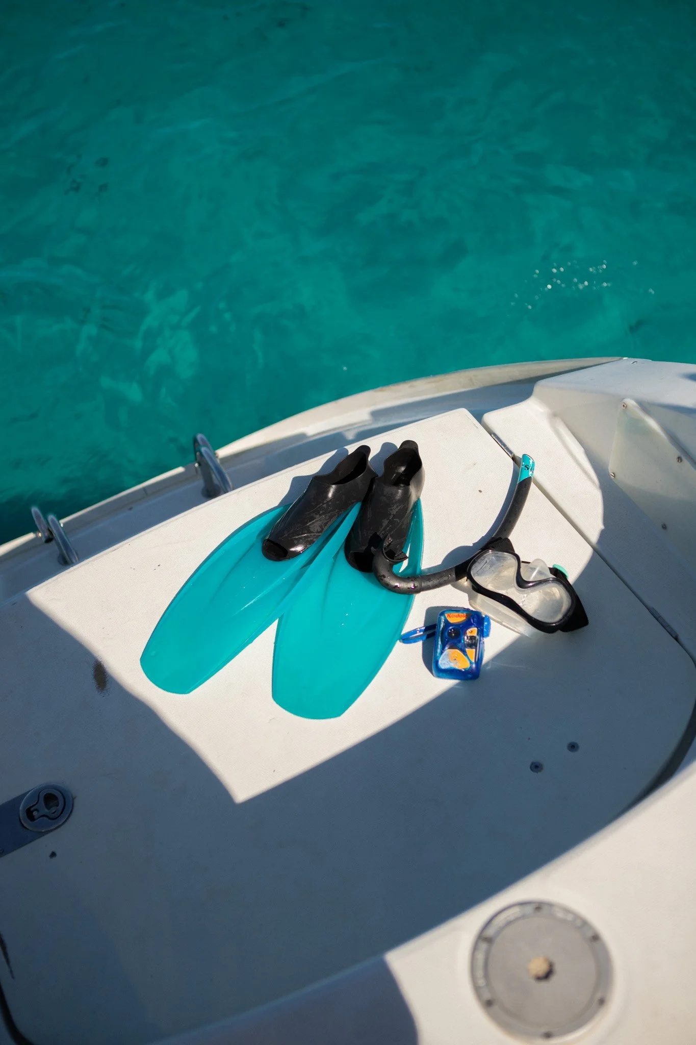 Zapatos de esnórquel, aletas de buceo, máscara y tubo de respiración, y una caja con juguetes de playa, en la cubierta de un barco, con agua clara y azul en el fondo.