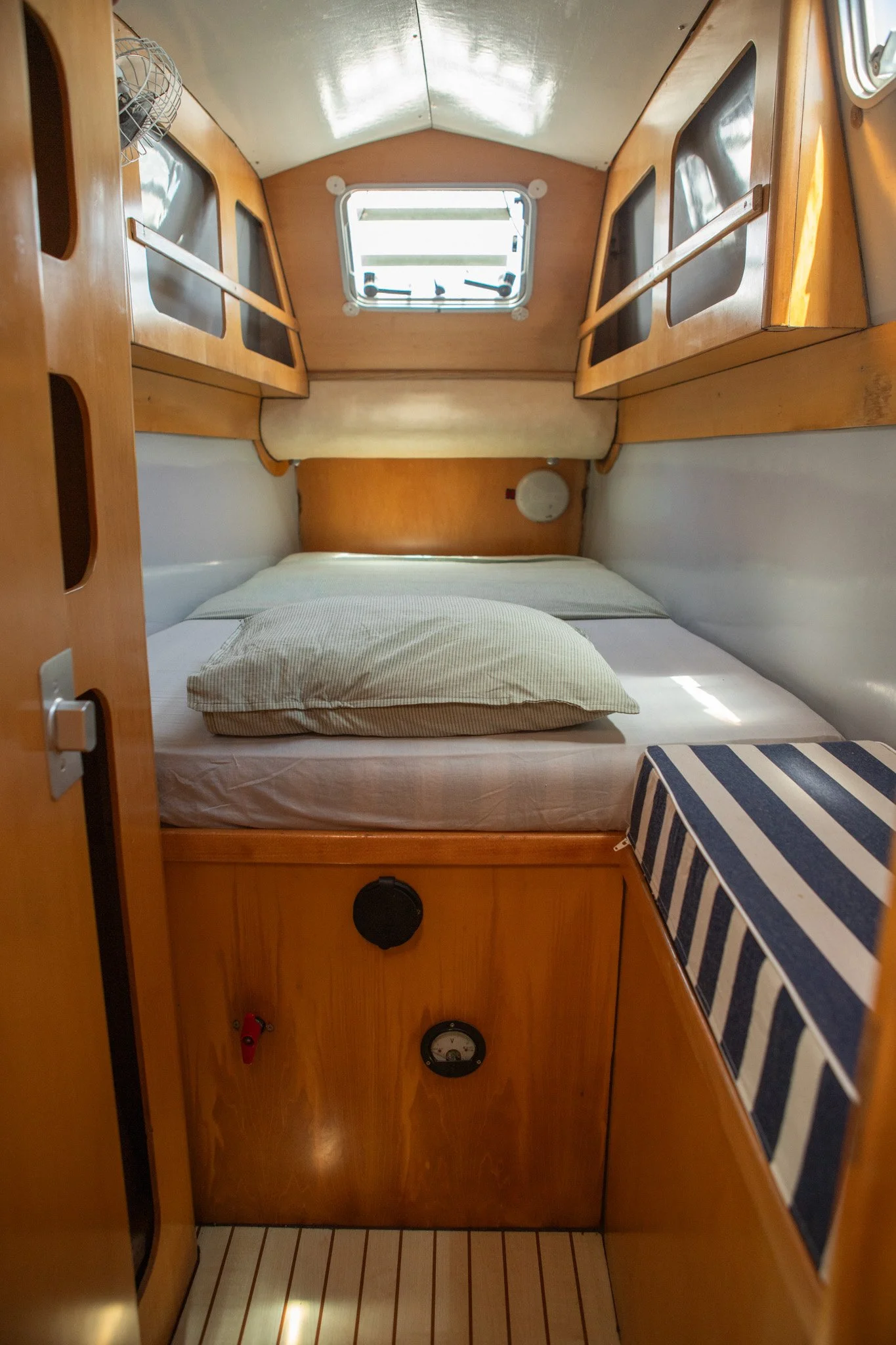 Interior de una cabina de barco con una cama con almohada, ventanas, armarios de madera, un panel con medidores y una pequeña plataforma con un tapete de rayas blancas y azules.