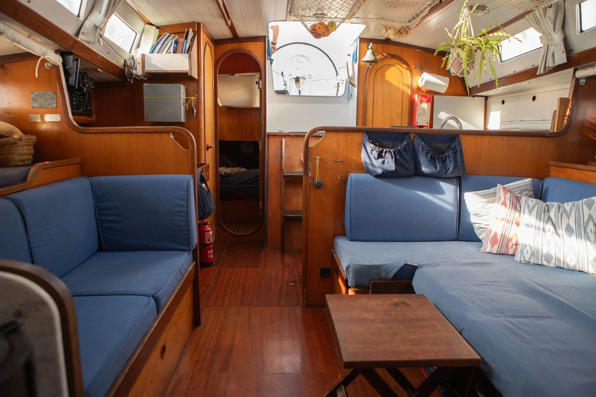 Interior de una embarcación de velero con asientos de madera y cojines azules, mostrando ventanas, plantas y objetos diversos, vistas al mar y la cabina.